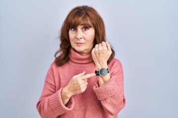 Middle age hispanic woman standing over isolated background in hurry pointing to watch time,...