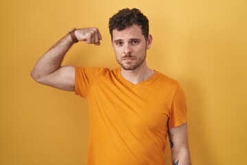 Young hispanic man standing over yellow background strong person showing arm muscle, confident and proud of power