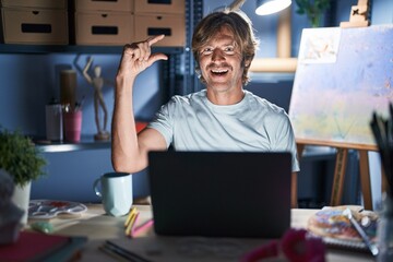 Middle age man sitting at art studio with laptop at night smiling and confident gesturing with hand doing small size sign with fingers looking and the camera. measure concept.