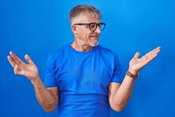 Naklejka premium Hispanic man with grey hair standing over blue background smiling showing both hands open palms, presenting and advertising comparison and balance