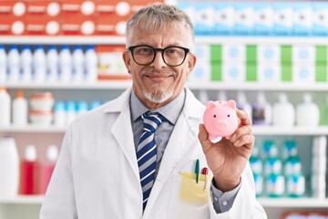 Hispanic man with grey hair working at pharmacy drugstore holding piggy bank looking positive and...