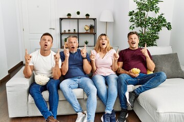 Group of middle age people sitting on the sofa at home amazed and surprised looking up and pointing with fingers and raised arms.
