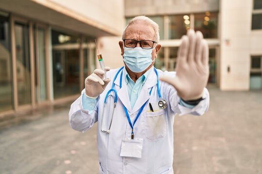 Senior Doctor With Grey Hair Wearing Safety Mask Holding Syringe With Open Hand Doing Stop Sign With Serious And Confident Expression, Defense Gesture