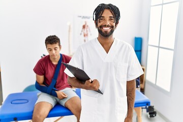 Young hispanic man working at pain recovery clinic with a man with broken arm with a happy and cool smile on face. lucky person.