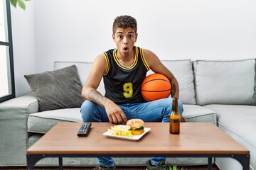 Young handsome hispanic man holding basketball ball cheering tv game scared and amazed with open mouth for surprise, disbelief face