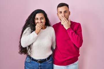 Young hispanic couple standing over pink background laughing and embarrassed giggle covering mouth...