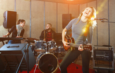 Attractive glad pleasant female soloist playing guitar and singing with her music band in sound studio