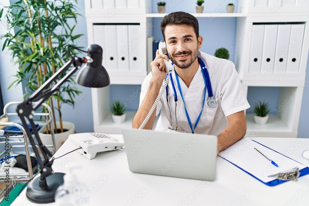 Wall mural Young hispanic man wearing doctor uniform talking on the telephone working at clinic