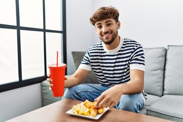 Young arab man smiling confident eating fried chicken drinking soda beverage at home