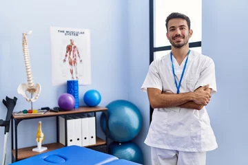 Fototapete Massagesalon Young hispanic man wearing physiotherapist uniform standing with arms crossed gesture at rehab clinic