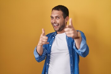 Hispanic man standing over yellow background pointing fingers to camera with happy and funny face. good energy and vibes.