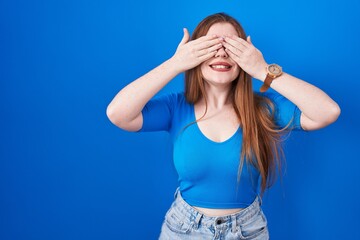 Redhead woman standing over blue background covering eyes with hands smiling cheerful and funny....
