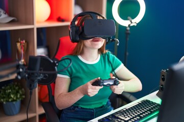 Fototapeta na wymiar Young redhead woman streamer playing video game using virtual reality glasses and joystick at gaming room