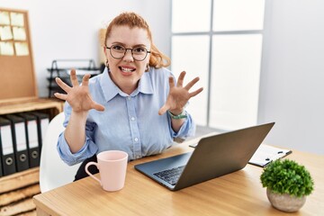 Young redhead woman working at the office using computer laptop smiling funny doing claw gesture as...