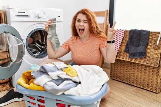 Young redhead woman putting dirty laundry into washing machine smiling with happy face winking at the camera doing victory sign with fingers. number two.