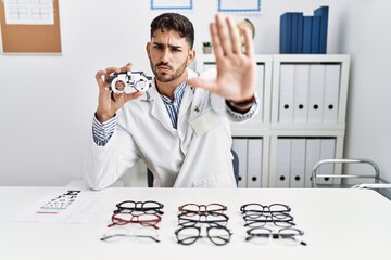 Young optician man holding optometry glasses doing stop sing with palm of the hand. warning...