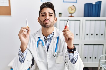 Young doctor man holding electronic cigarette at medical clinic looking at the camera blowing a kiss being lovely and sexy. love expression.