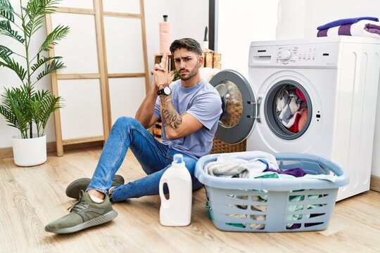 Young hispanic man putting dirty laundry into washing machine holding symbolic gun with hand gesture, playing killing shooting weapons, angry face