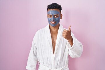 Young hispanic man wearing beauty face mask and bath robe doing happy thumbs up gesture with hand....