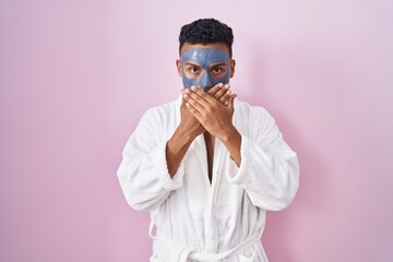 Young hispanic man wearing beauty face mask and bath robe shocked covering mouth with hands for mistake. secret concept.