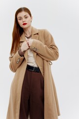 Shooting for the outerwear catalog. Red-haired beauty in a beige full-length coat in the studio on a white background
