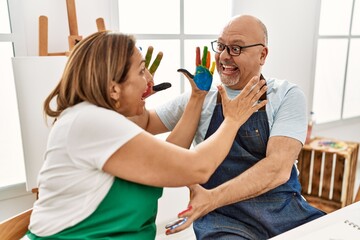 Middle age hispanic painter couple smiling happy touching face with painted hands at art studio.