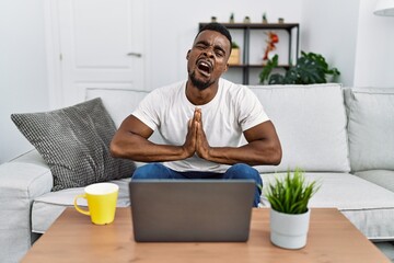 Young african man using laptop at home begging and praying with hands together with hope expression...