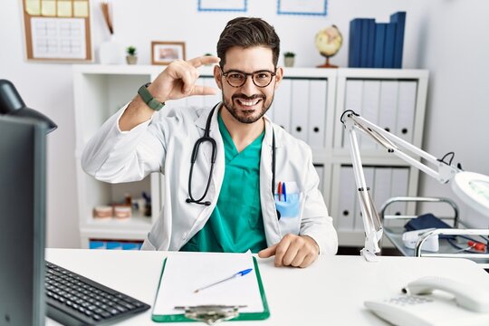 Young man with beard wearing doctor uniform and stethoscope at the clinic smiling and confident gesturing with hand doing small size sign with fingers looking and the camera. measure concept.