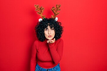 Young middle east woman wearing cute christmas reindeer horns looking at the camera blowing a kiss with hand on air being lovely and sexy. love expression.