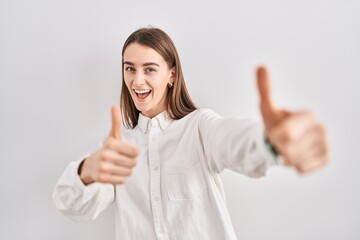 Young caucasian woman standing over isolated background approving doing positive gesture with hand, thumbs up smiling and happy for success. winner gesture.