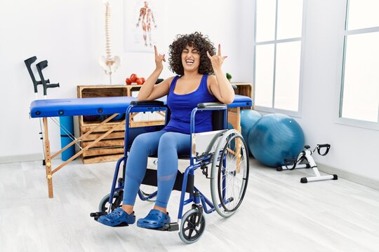 Young middle eastern woman sitting on wheelchair at physiotherapy clinic shouting with crazy expression doing rock symbol with hands up. music star. heavy concept.
