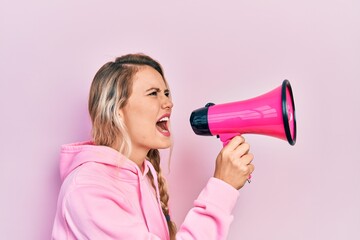 Young beautiful blonde caucasian woman shouthing and screaming through megaphone over isolated pink background