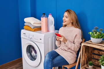 Young woman using smartphone waiting for washing machine at laundry room