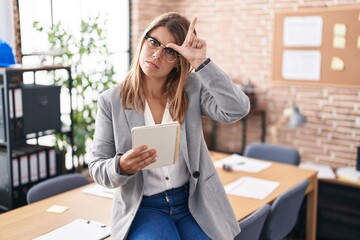 Young hispanic woman working at the office wearing glasses making fun of people with fingers on...