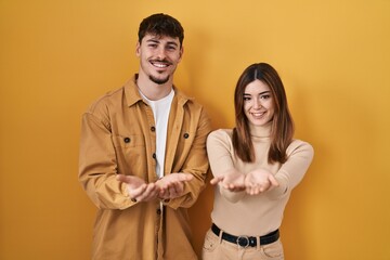 Young hispanic couple standing over yellow background smiling with hands palms together receiving or giving gesture. hold and protection