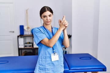 Young hispanic woman wearing physiotherapist uniform standing at clinic holding symbolic gun with hand gesture, playing killing shooting weapons, angry face