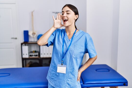 Young hispanic woman wearing physiotherapist uniform standing at clinic shouting and screaming loud to side with hand on mouth. communication concept.