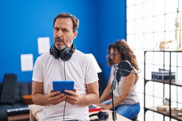 Middle age hispanic man at music studio using tablet in shock face, looking skeptical and...