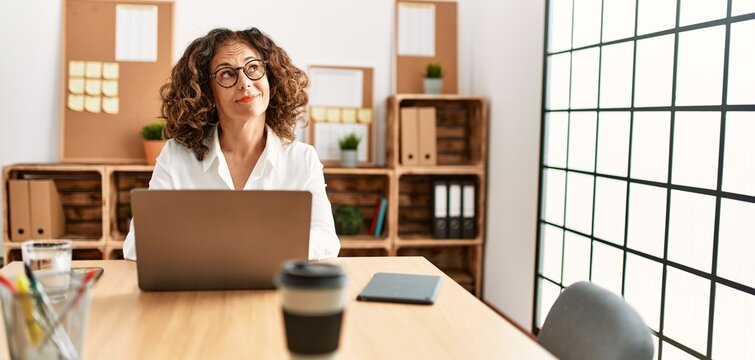 Middle age hispanic woman working at the office wearing glasses smiling looking to the side and staring away thinking.
