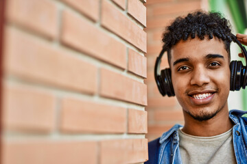 Hispanic young man listening to music wearing headphones leaning on bricks wall
