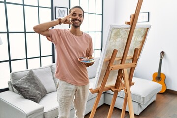 Young hispanic man with beard painting on canvas at home doing peace symbol with fingers over face, smiling cheerful showing victory