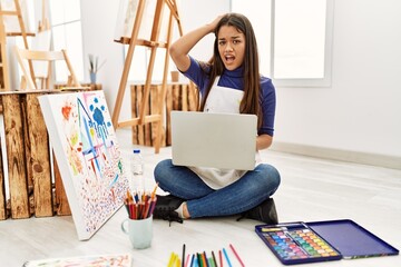 Young brunette woman sitting on the floor at art studio with laptop stressed and frustrated with...