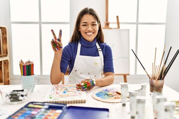 Young brunette woman at art studio with painted hands showing and pointing up with fingers number two while smiling confident and happy.