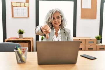 Middle age businesswoman sitting on desk working using laptop at office looking unhappy and angry showing rejection and negative with thumbs down gesture. bad expression.