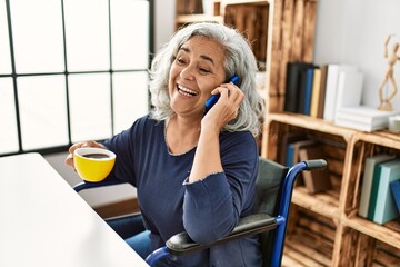 Middle age grey-haired disabled woman talking on the smartphone and drinking coffee sitting on...