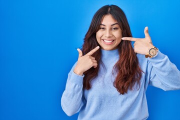 Fototapeta premium Hispanic young woman standing over blue background smiling cheerful showing and pointing with fingers teeth and mouth. dental health concept.