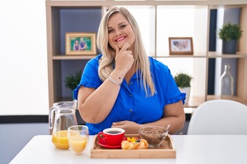 Caucasian plus size woman eating breakfast at home looking confident at the camera smiling with crossed arms and hand raised on chin. thinking positive.