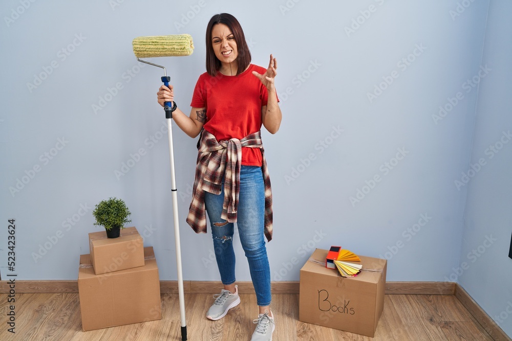 Sticker Young hispanic woman painting home walls with paint roller crazy and mad shouting and yelling with aggressive expression and arms raised. frustration concept.