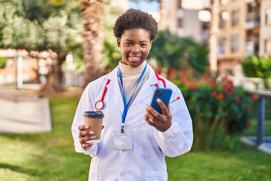African american woman wearing doctor uniform using smartphone drinking coffee at park