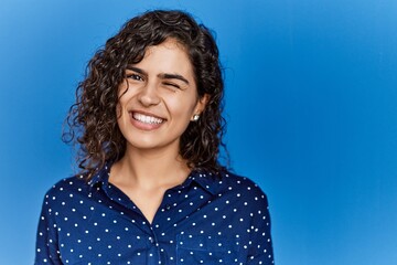 Young brunette woman with curly hair wearing casual clothes over blue background winking looking at the camera with sexy expression, cheerful and happy face.